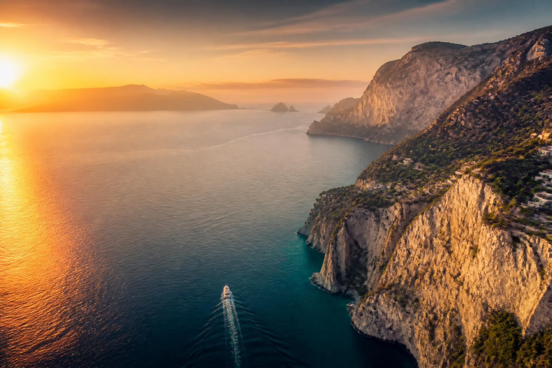 Aerial view of a Mediterranean coastline at golden hour with a single yacht on open water